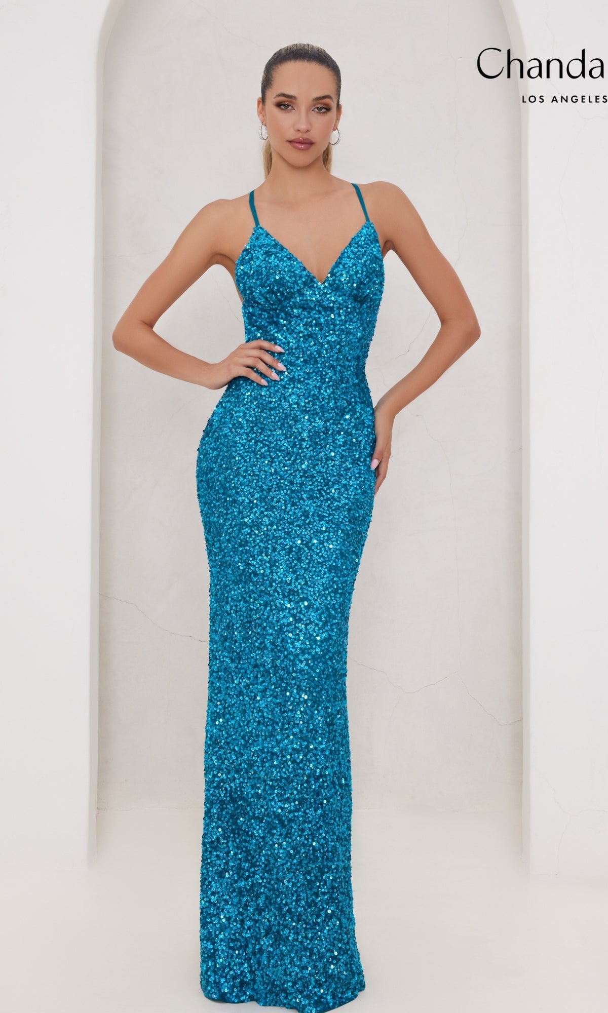 A woman poses in the Long Prom Dress 30045 by Chandalier, a fitted blue sequin gown with thin straps and a v-neckline, its floor-length silhouette shimmering against a plain light background.